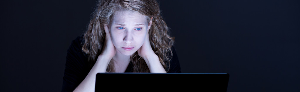 A sad girl looks at a glowing computer screen in the midst of a dark room.