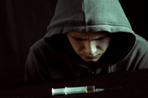 A young man in a hoodie looks at a needle on a black table, tortured by his addiction.